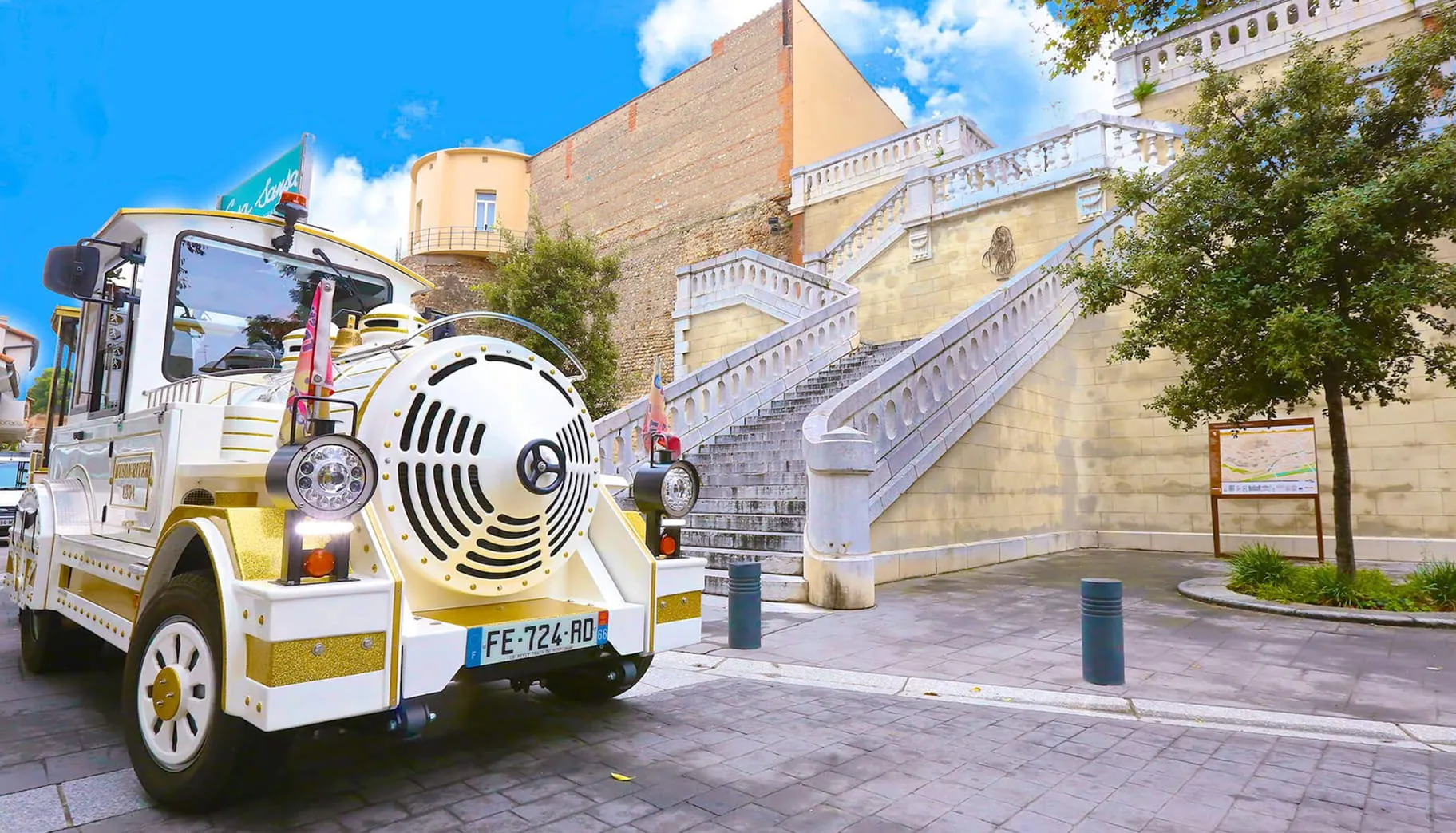 Pequeño tren turístico blanco y dorado aparcado junto a una escalinata monumental de piedra bajo un cielo azul parcialmente nublado.