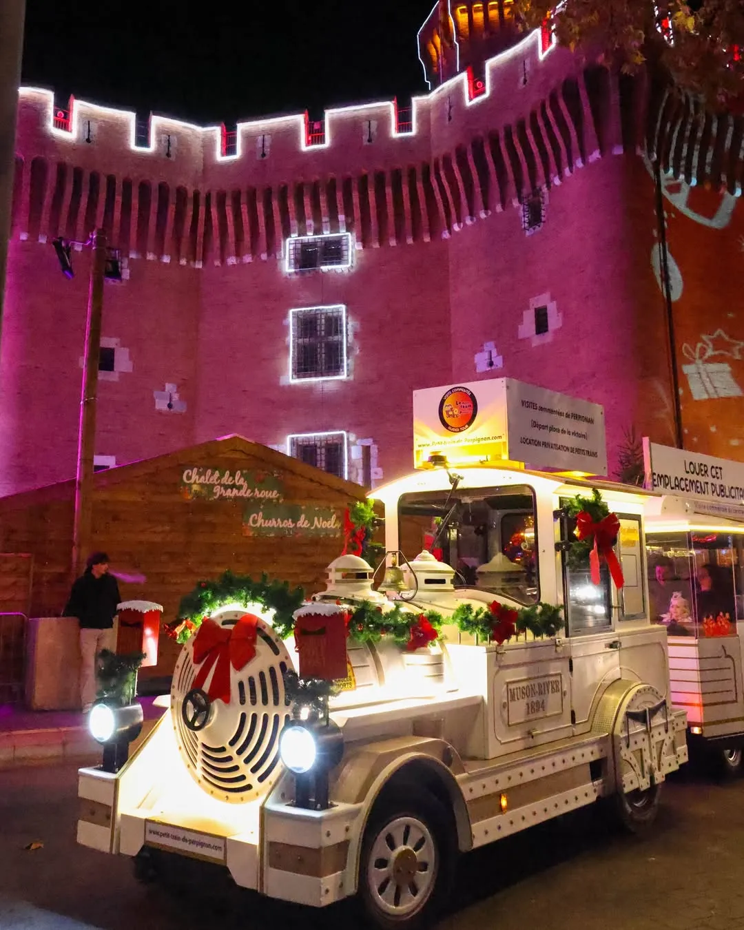Pequeño tren blanco decorado con guirnaldas navideñas y lazos rojos frente a un castillo iluminado en morado por la noche.
