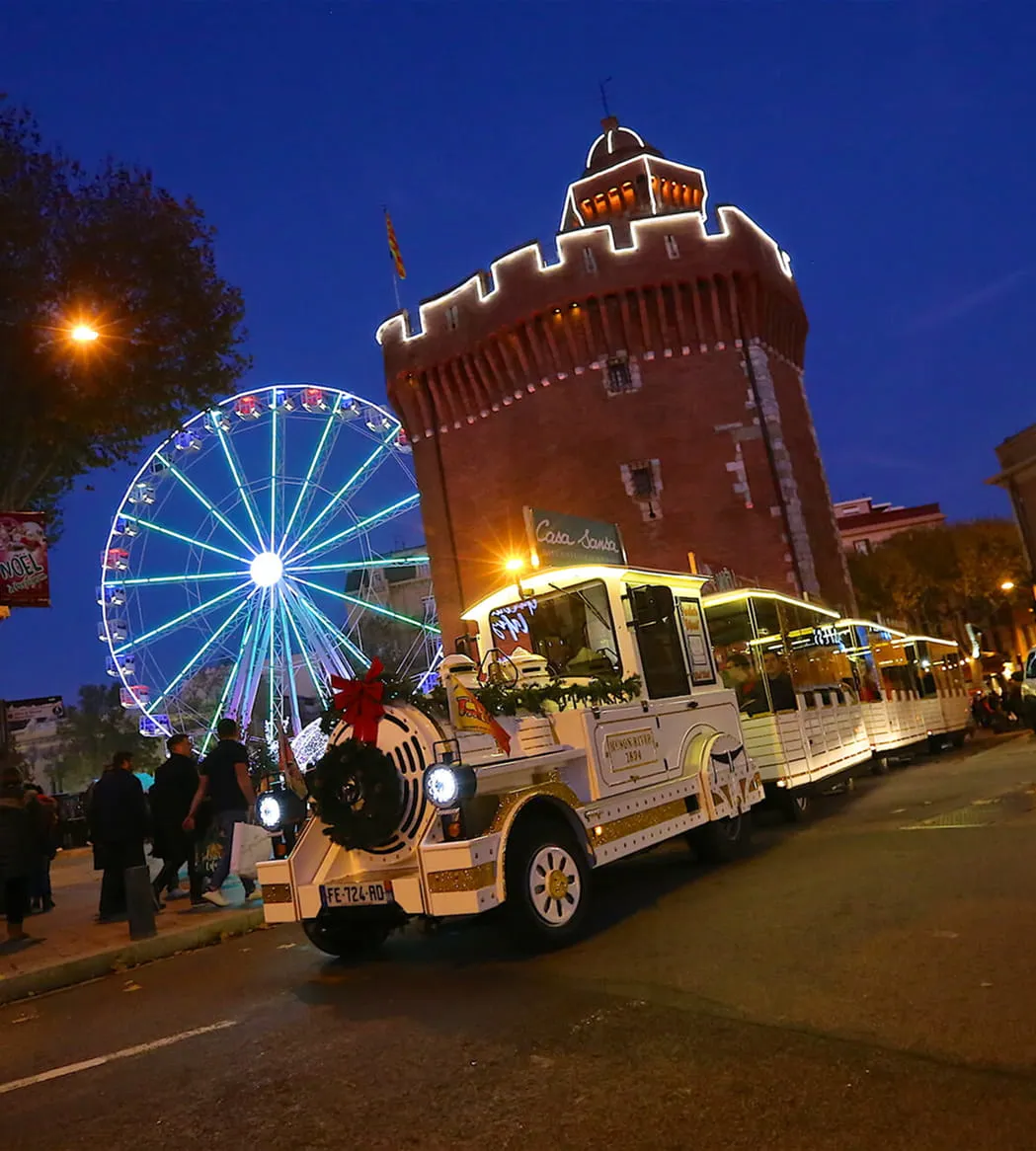 Tren turístico blanco iluminado decorado para Navidad pasando frente a una noria luminosa y una torre histórica al atardecer.