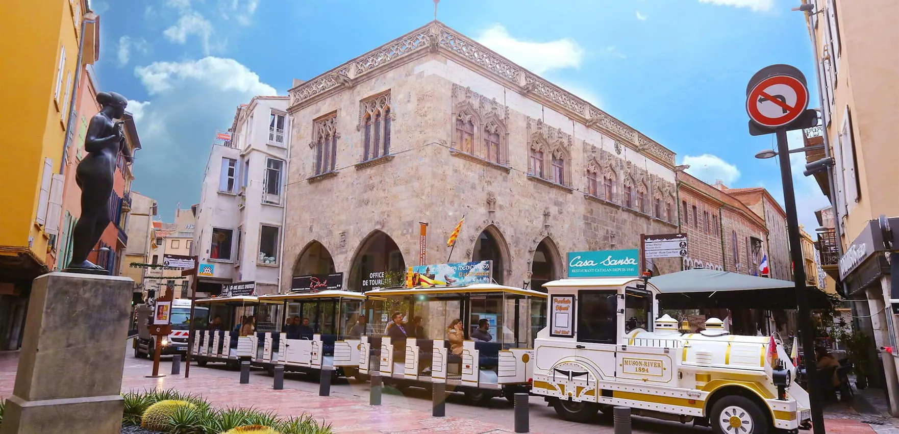 White tourist train with passengers driving in front of a historic stone building with gothic windows, a black statue on a pedestal on the left, and blue sky with a few clouds.