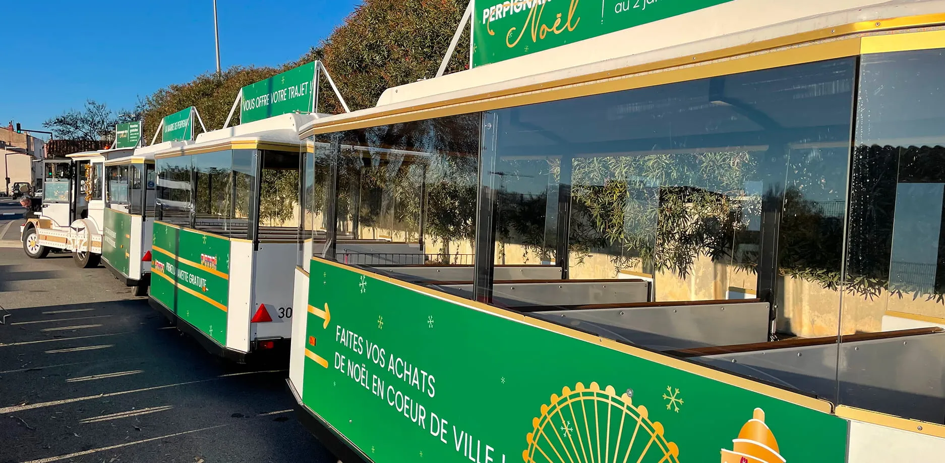 White and green tourist train parked on a road with green advertising panels for downtown Christmas shopping.