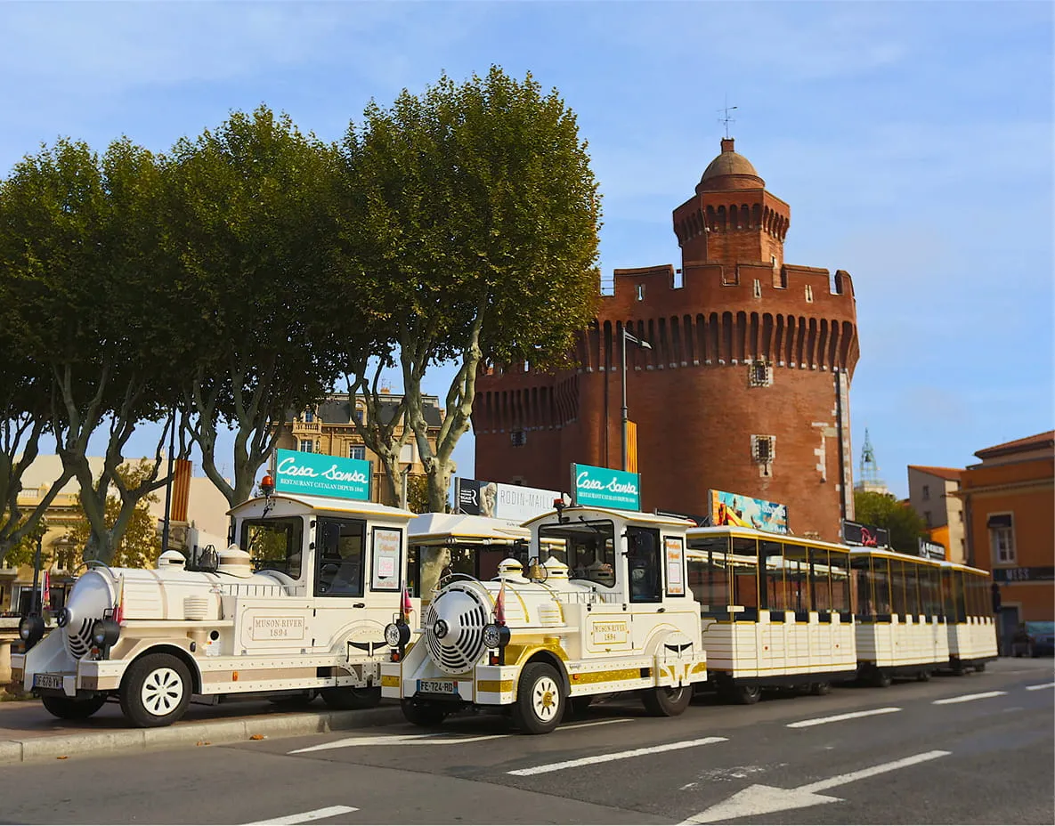 White tourist train parked in front of a red brick tower surrounded by green trees.