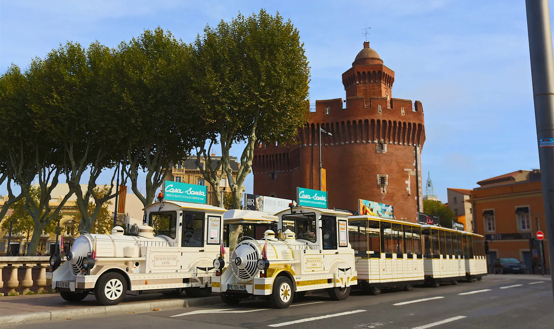 Pequeño tren turístico blanco aparcado junto a un edificio de ladrillo rojo en forma de torre, con árboles frondosos cerca.