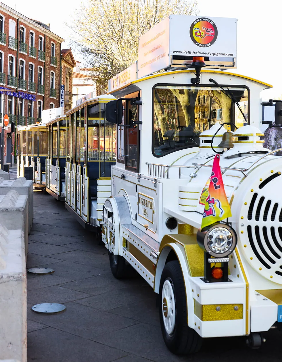 Pequeño tren turístico blanco y dorado aparcado a lo largo de una calle adoquinada con edificios al fondo en Perpiñán.