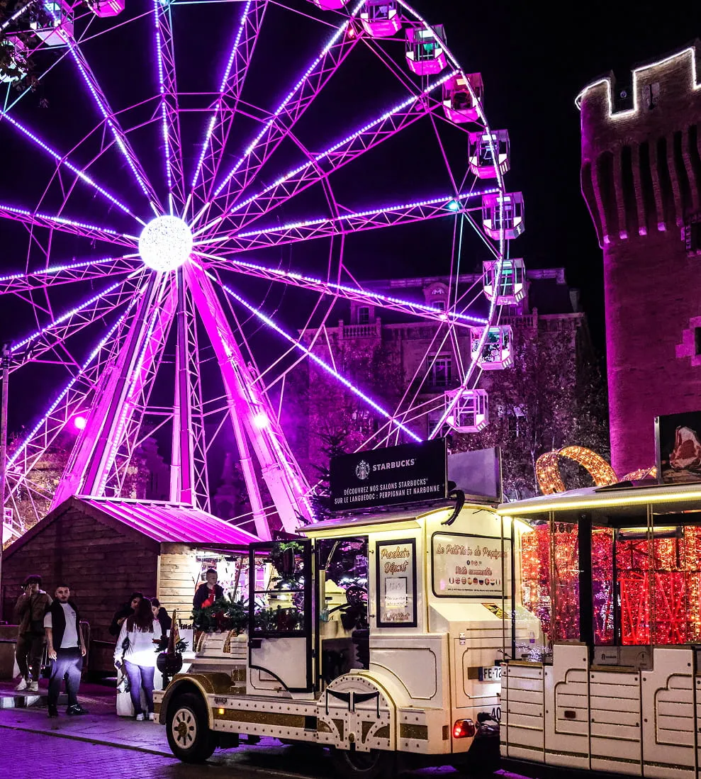 Noria iluminada en morado por la noche junto a un pequeño tren turístico blanco y una antigua torre iluminada.